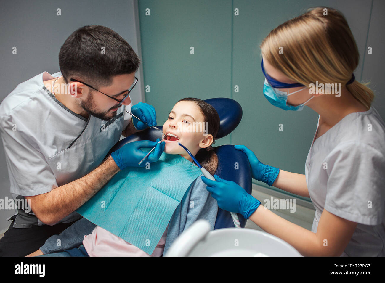Male dentist check up girl's teeth with dentist's tools. Female helper stand beside. Girl sit in dental chair in room. She keep mouth opened Stock Photo