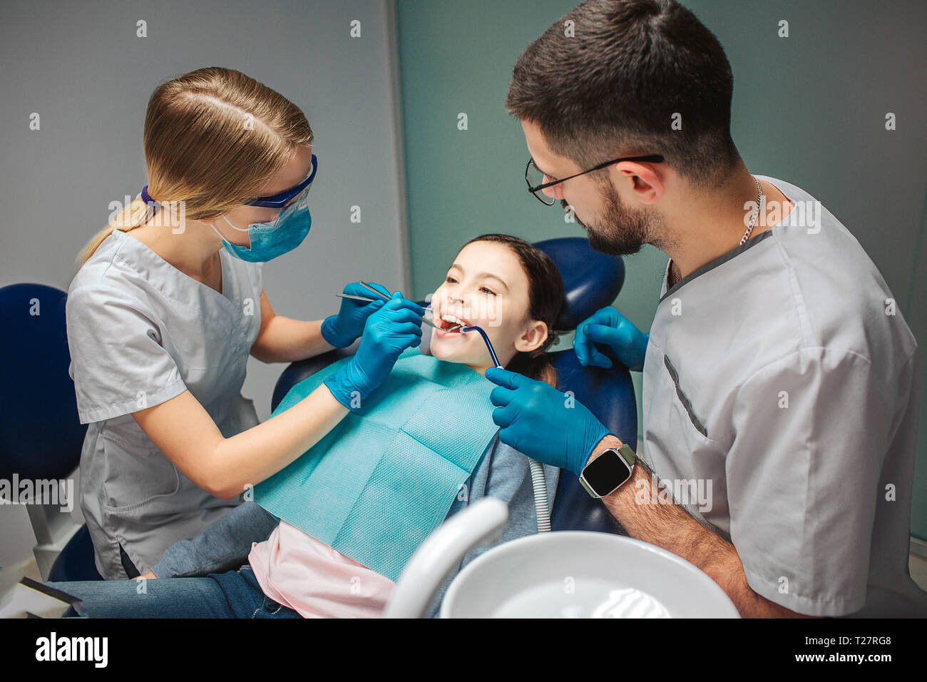 Female dentist check up girl's teeth. Male helper stad beside. Girl sit in dental chair in room. She keep mouth opened Stock Photo