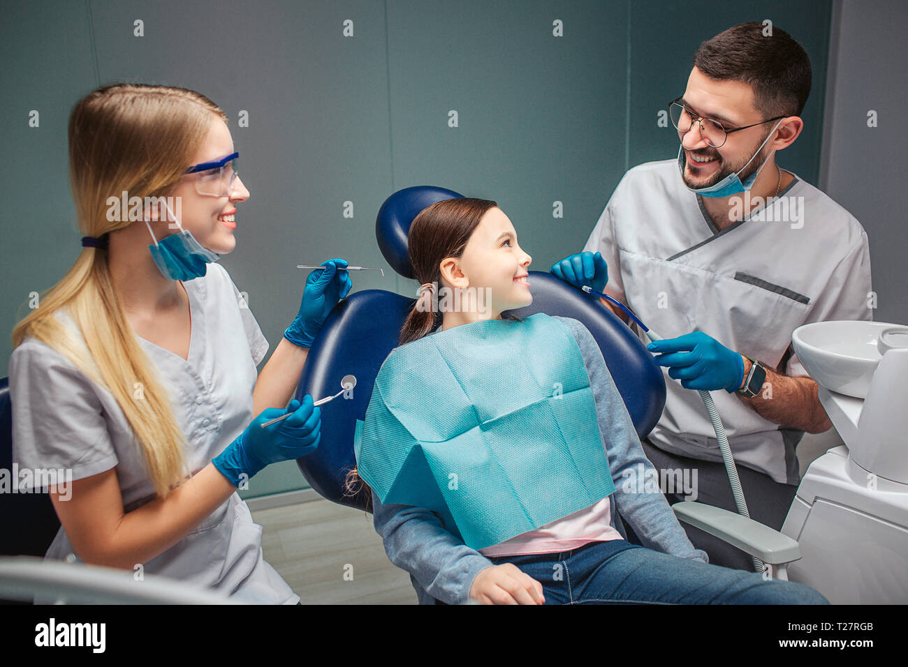 Positive happy girl sit in dental chair and look and male dentist. He smile to her. Female helper happy too. Sit in room together Stock Photo