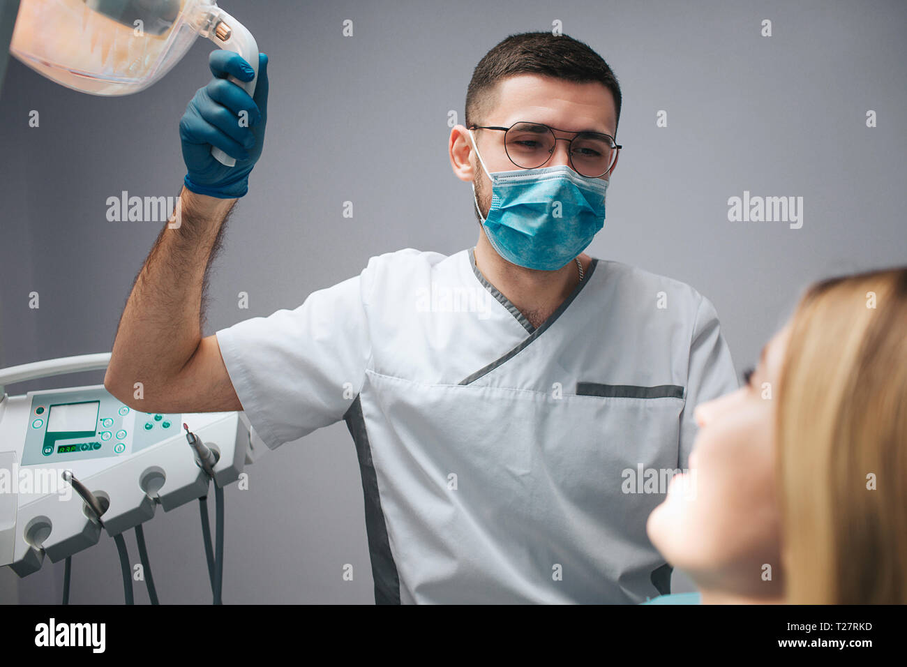 Young dentist in mask sit at client and hold lamp. Young woman look at doctor and smile. They