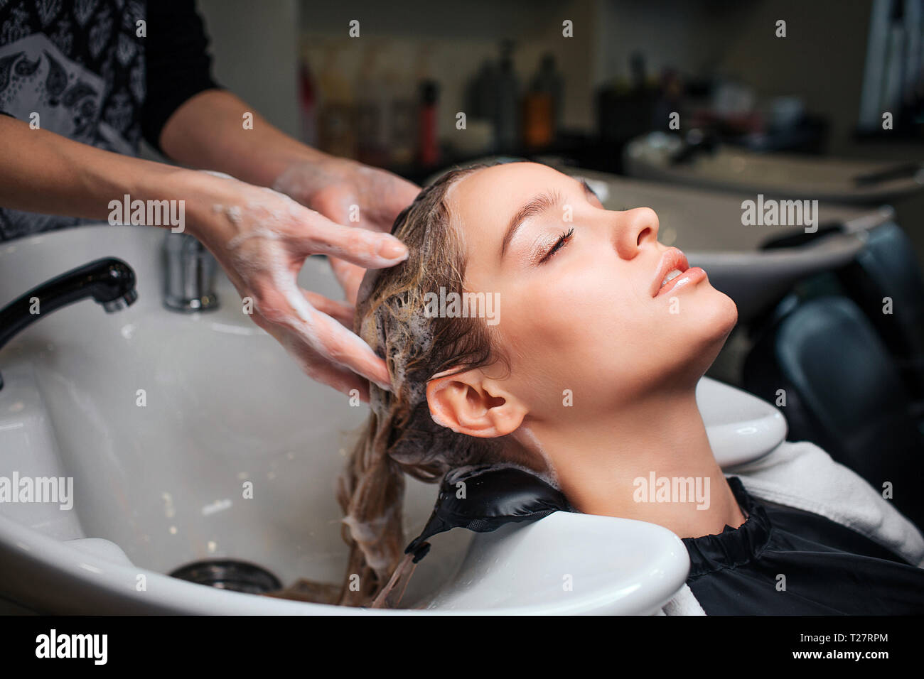 Beautiful young woman sitting near sink while hairdresser washing her hair in beauty salon. Hair stylist at work Stock Photo