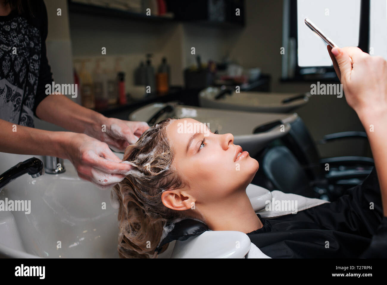 Beautiful young woman looking at phone while hairdresser washing her hair after mask in beauty salon. Beauty and people concept Stock Photo