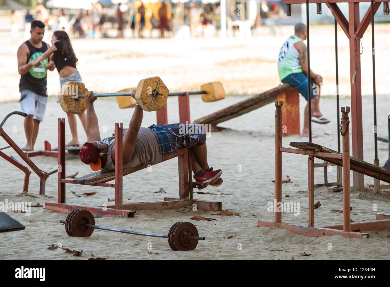 A man works out doing bench press with crude weightlifting equipment in an outdoor gym on a public beach on July 8, 2018 in Sao Vicente, Brazil. Stock Photo