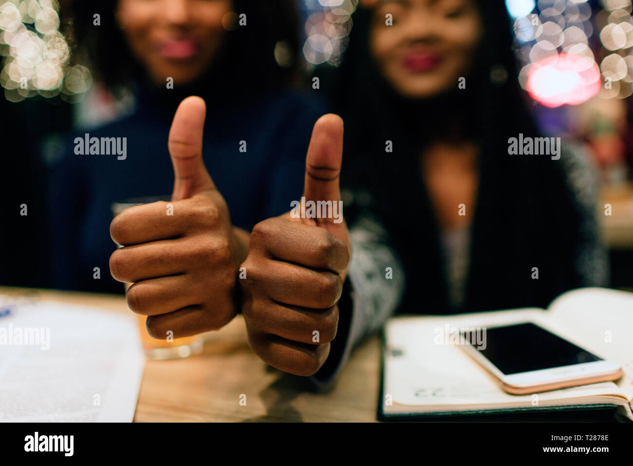 Close up of two hands hold big thumb up. Young african women smile. They have study materials and phone at table Stock Photo