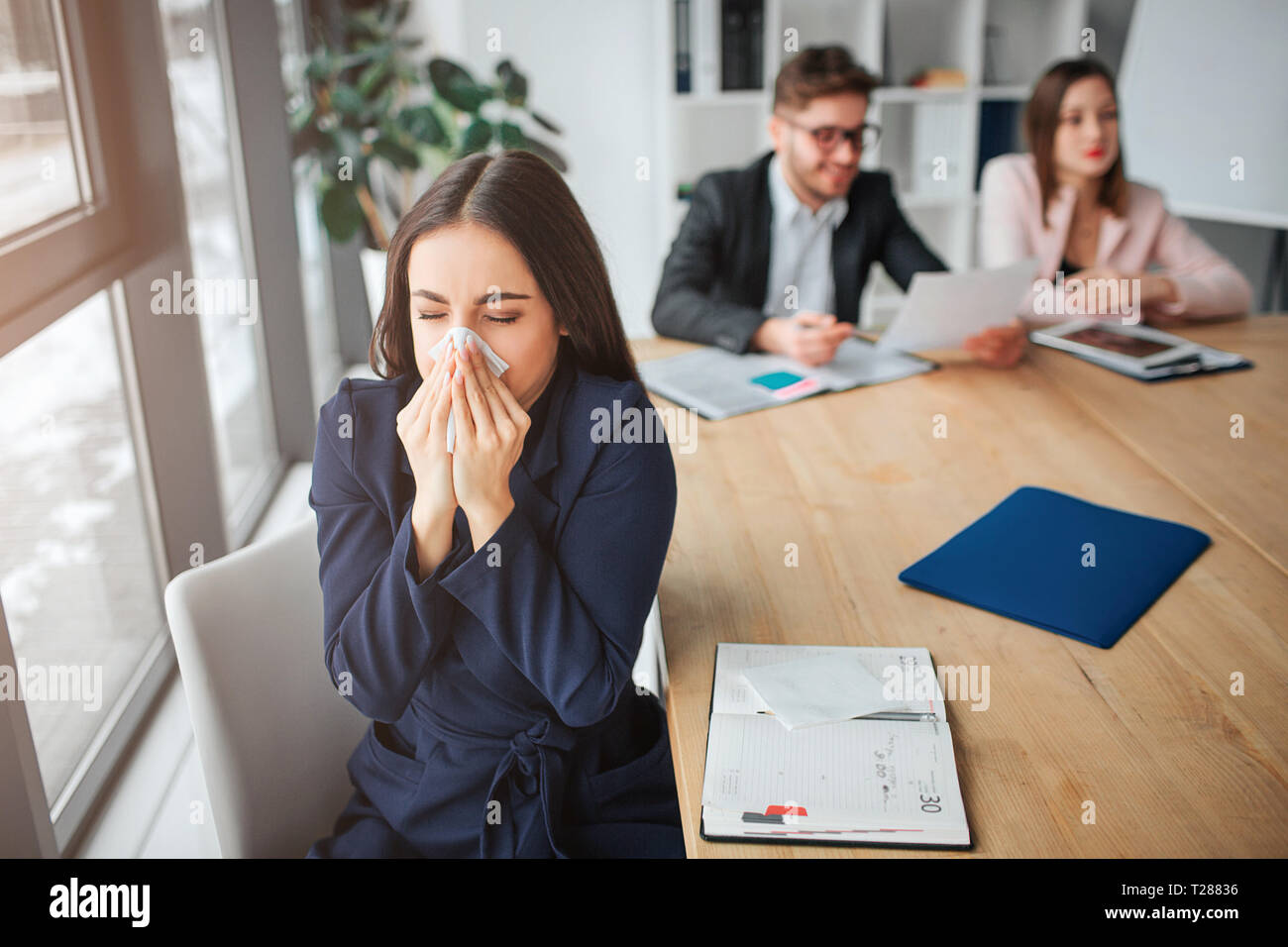 Sick young woman sit saparately at table in meeting room and sneezing. She hold white napkin. Her colleagues work behind Stock Photo