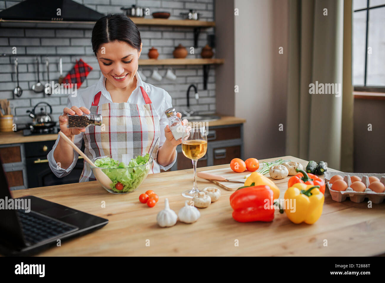 Cheerful nice woman sit at table in kitchen and put spices in bowl with salad. She looks happy. Woman wears apron. Colorful vegetables and eggs stand  Stock Photo