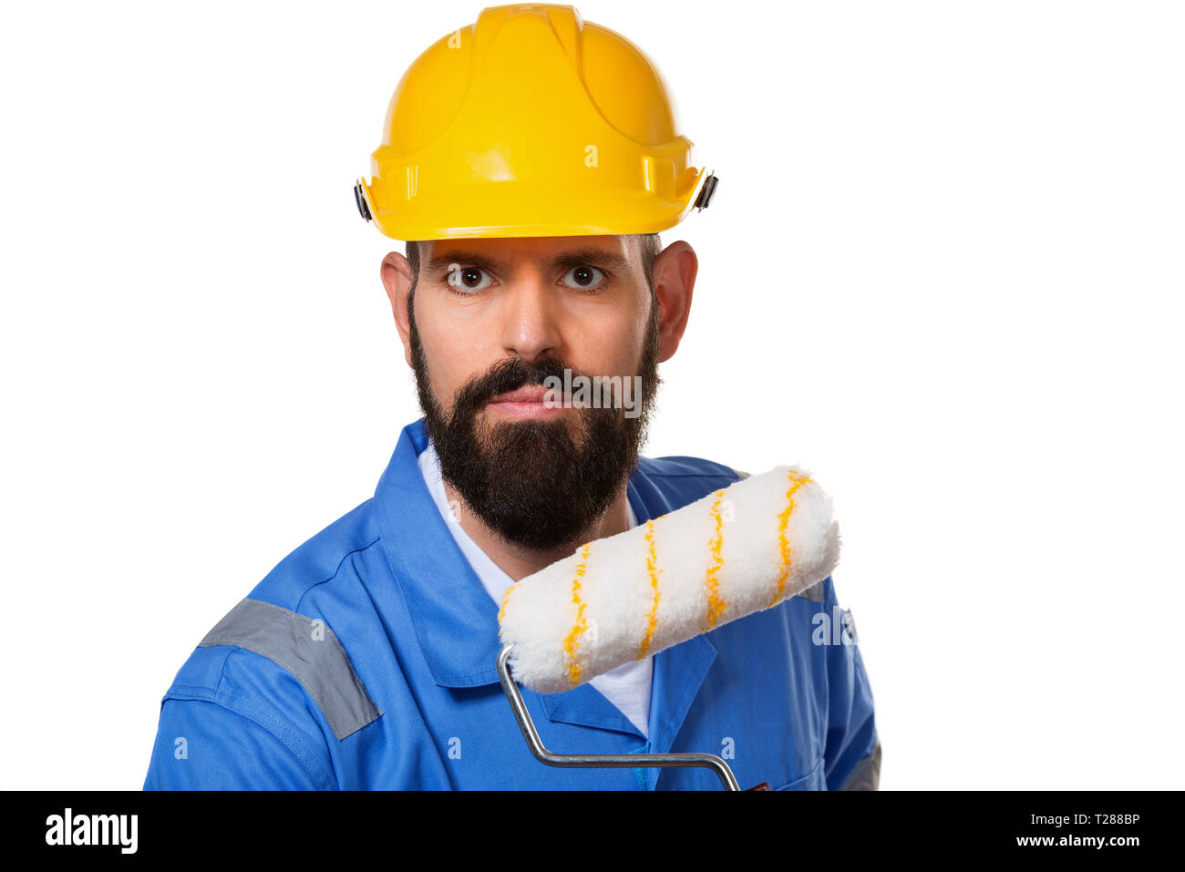 Bearded builder in yellow helmet and blue uniform holding paint roller. Isolated on white background. Stock Photo