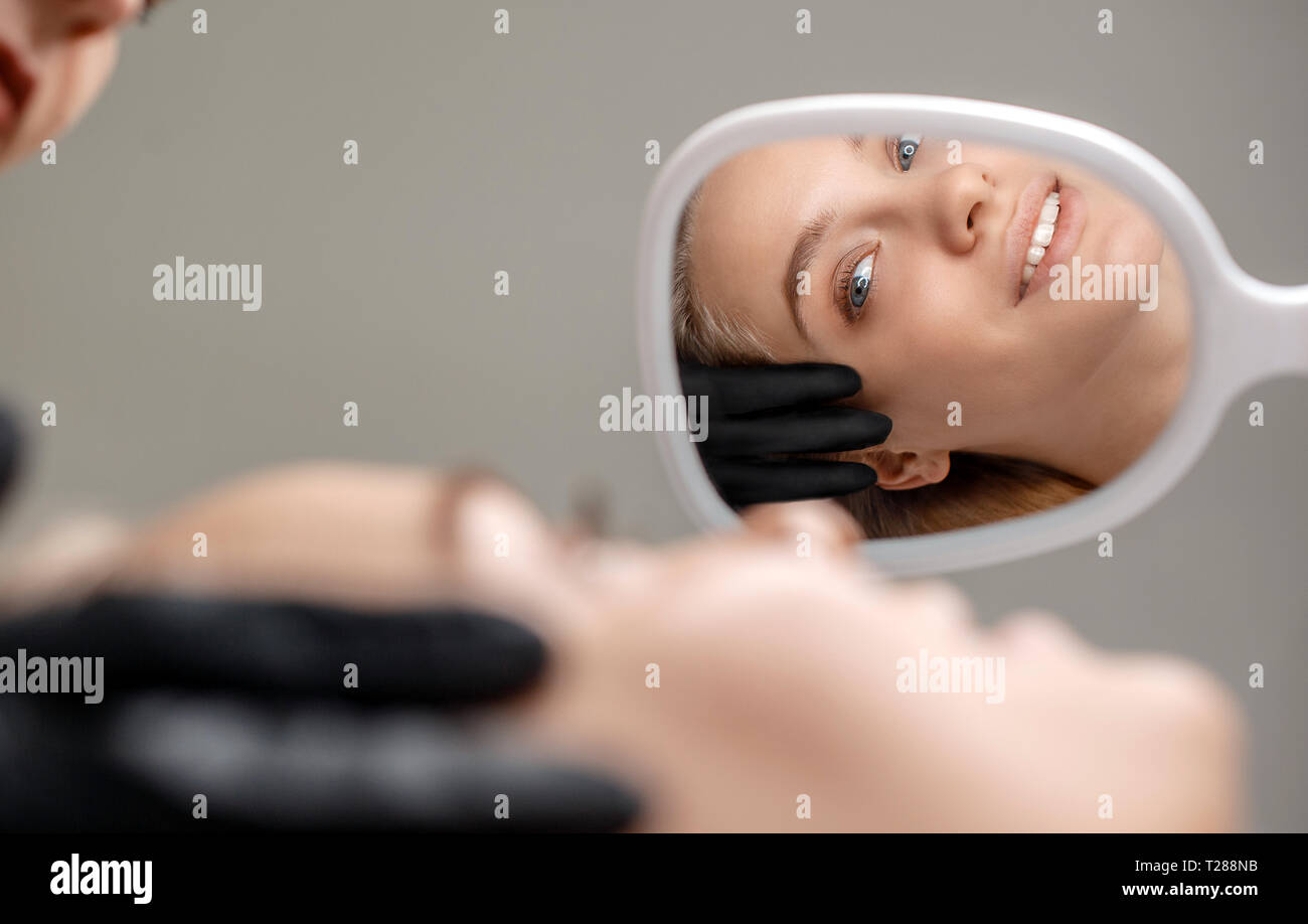 Young woman lying on beautician's couch and look in miroor. She smile ...