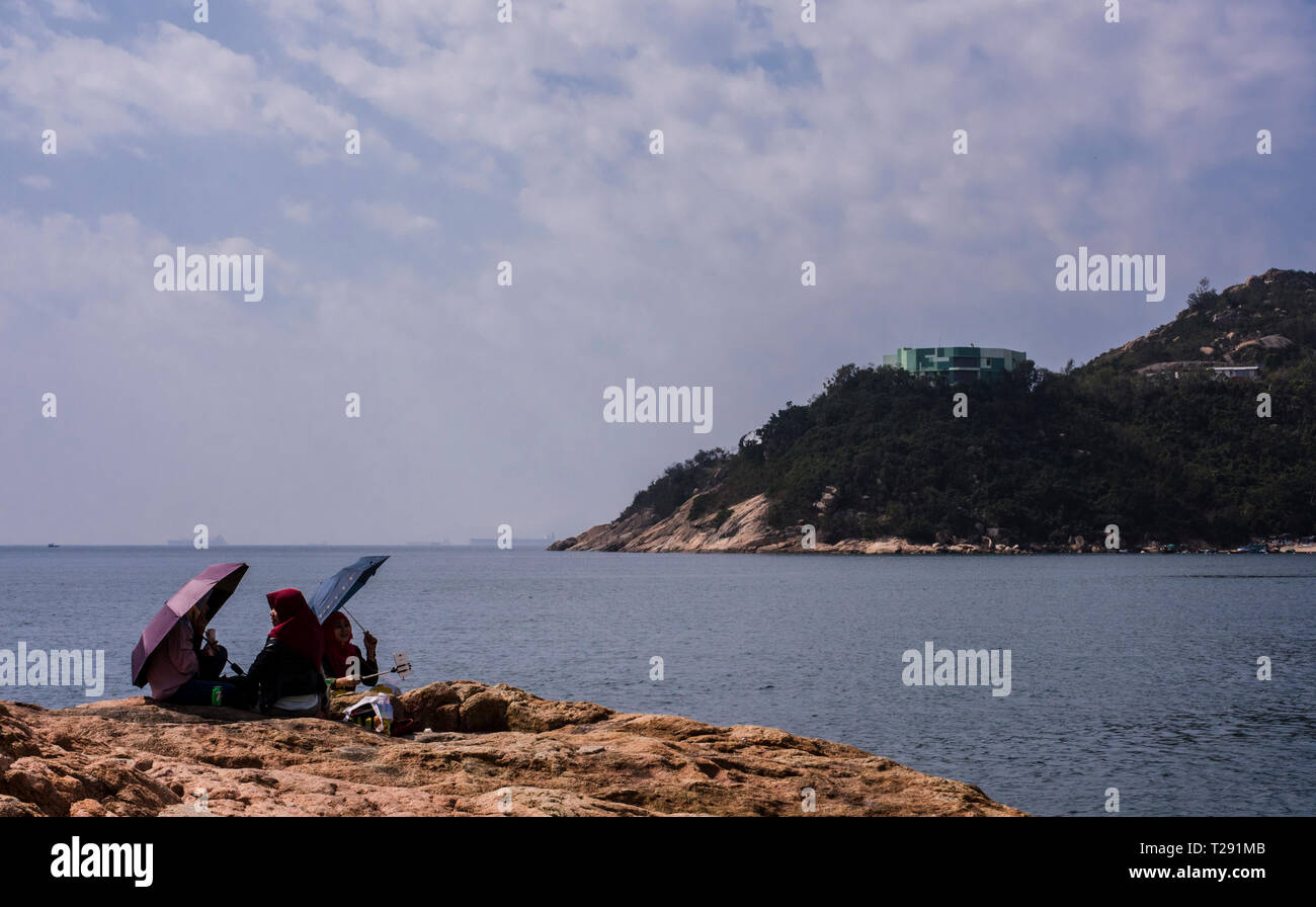 Three people sitting on rocks, using umbrellas to shield themselves from the sun, in the seaside village of Stanley, Hong Kong Stock Photo