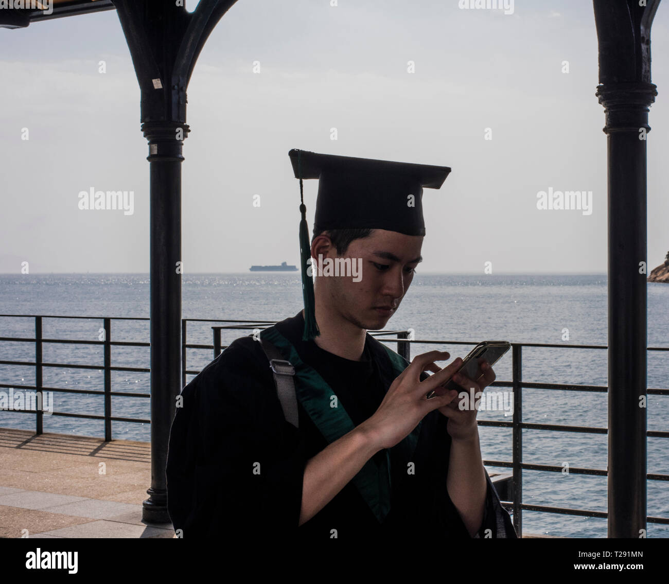 Male student, wearing graduation gown, using smartphone, in the seaside