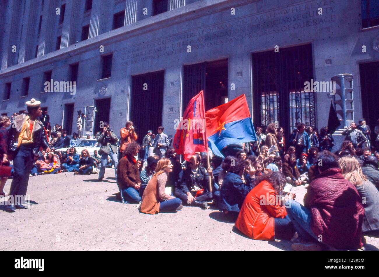 During the 1971 May Day Protests against the Vietnam War, a large circle of warmly dressed male and female protestors sit together outside the US Department of Justice building (now the Robert F Kennedy Department of Justice Building) holding two flags, including a red, blue, and gold Viet Cong Flag, in Washington, DC, May 1971. () Stock Photo