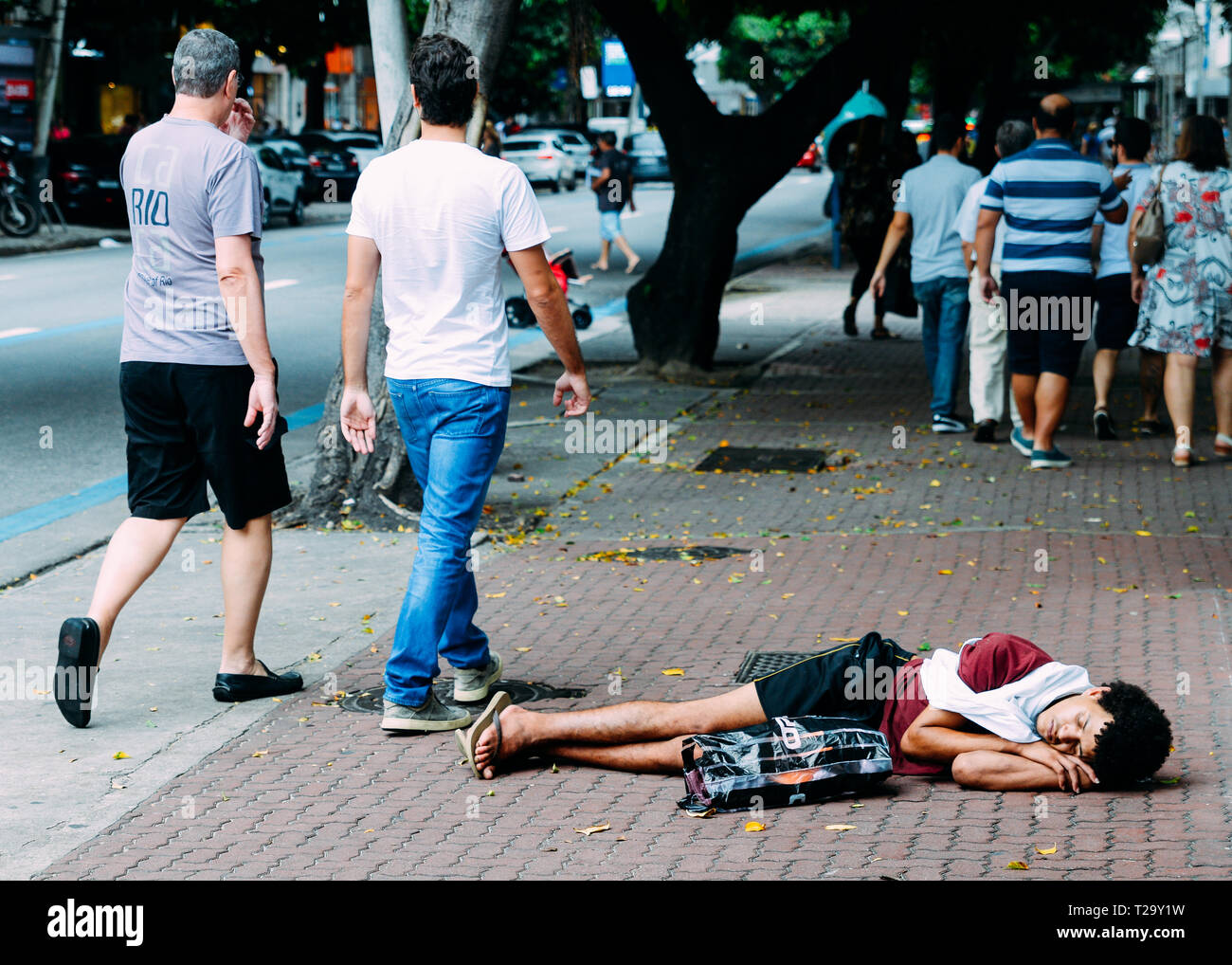 Rio de Janeiro, Brazil, March 23, 2019: Homeless young man sleeping rough while pedestrians walk next to him on busy pedestrian sidewalk in the wealth Stock Photo