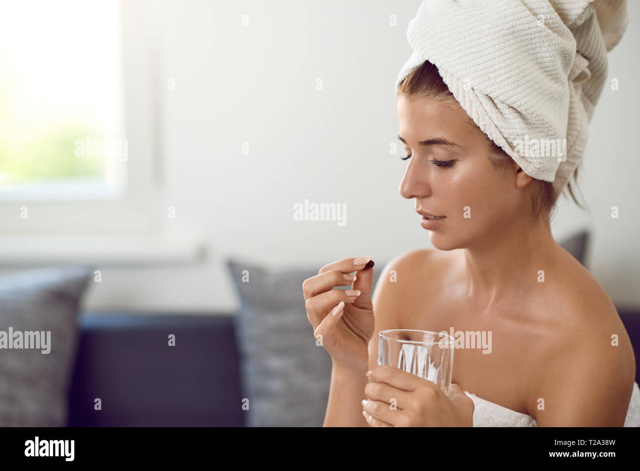 Attractive young woman with her hair and body wrapped in white towels after bathing or spa treatment taking a tablet or dietary supplement holding a g Stock Photo