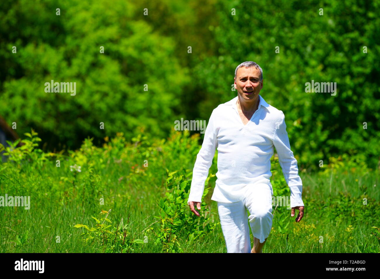 A man in white clothes walking in a field and touching flowers, surrounded by green. Stock Photo