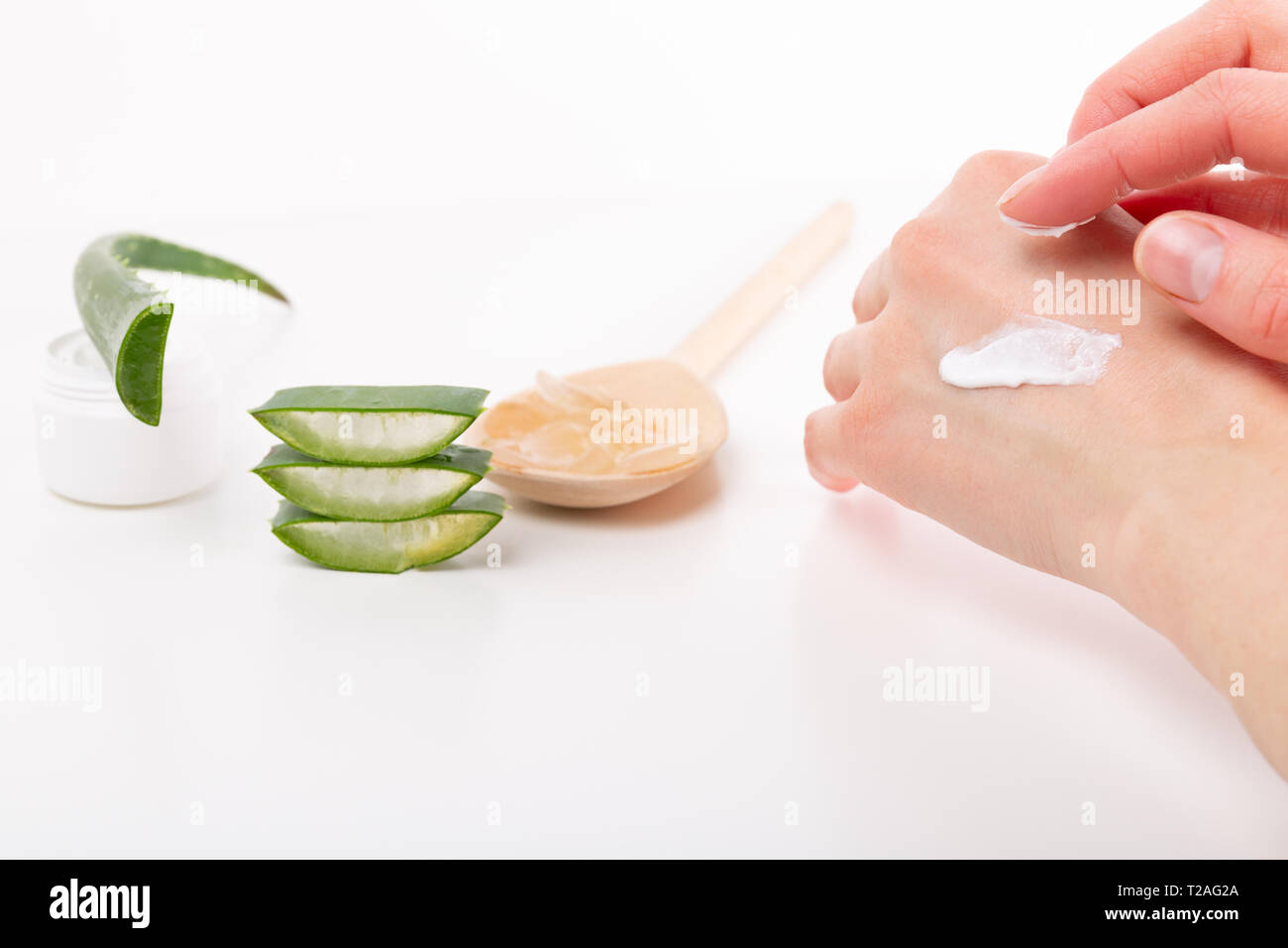 Opened plastic container with cream, aloe vera gel on woden spoon, sliced aloe vera plant and woman's hands on a white background Stock Photo