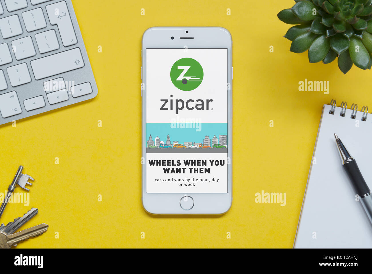 An iPhone showing the Zipcar website rests on a yellow background table with a keyboard, keys, notepad and plant (Editorial use only). Stock Photo