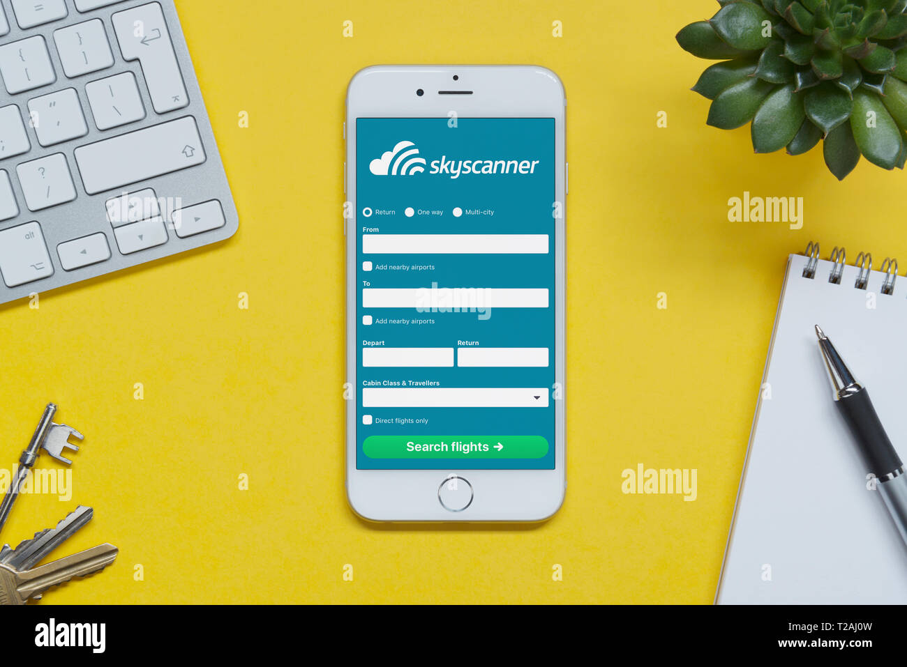 An iPhone showing the Skyscanner website rests on a yellow background table with a keyboard, keys, notepad and plant (Editorial use only). Stock Photo