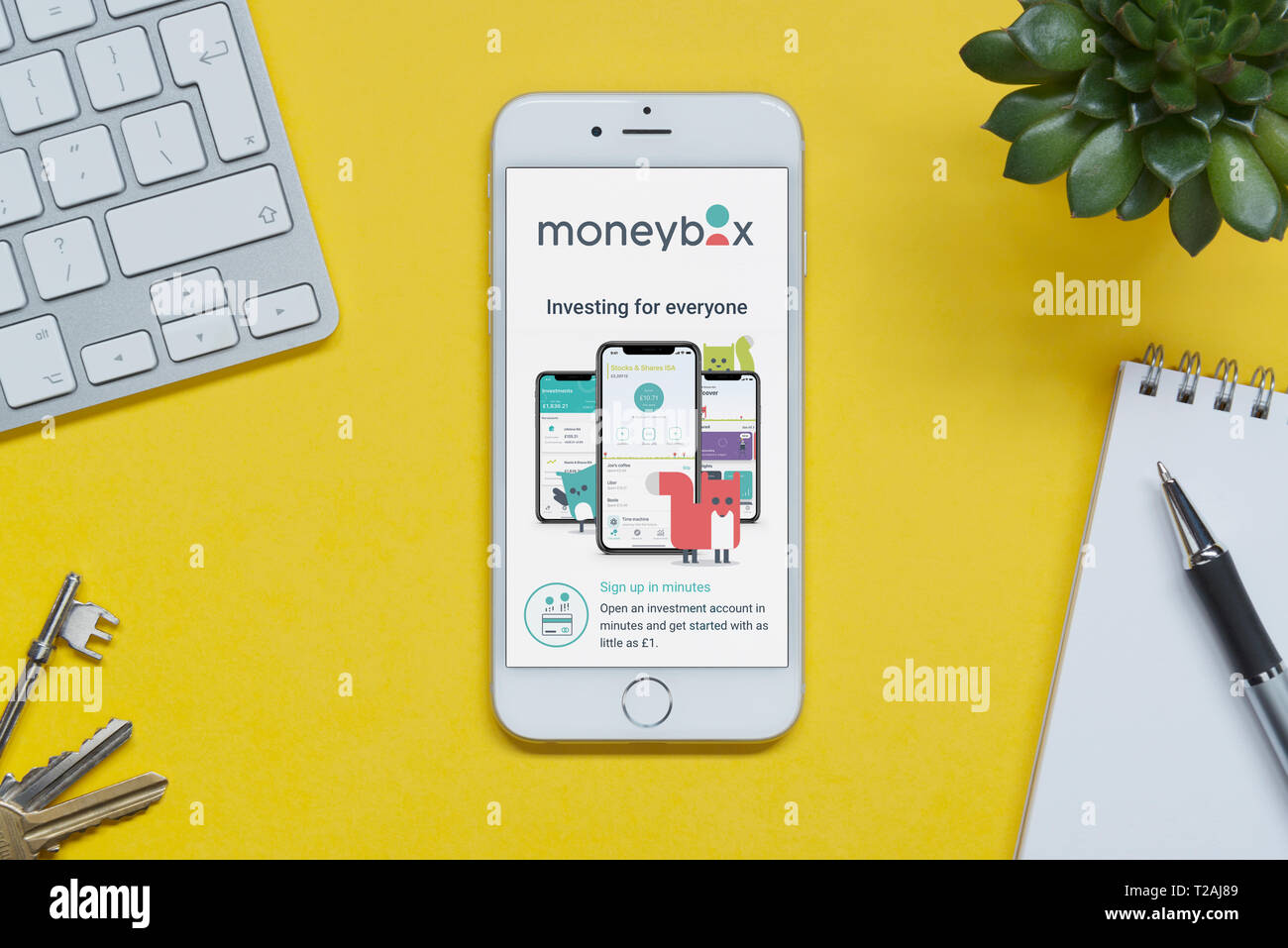 An iPhone showing the Moneybox website rests on a yellow background table with a keyboard, keys, notepad and plant (Editorial use only). Stock Photo