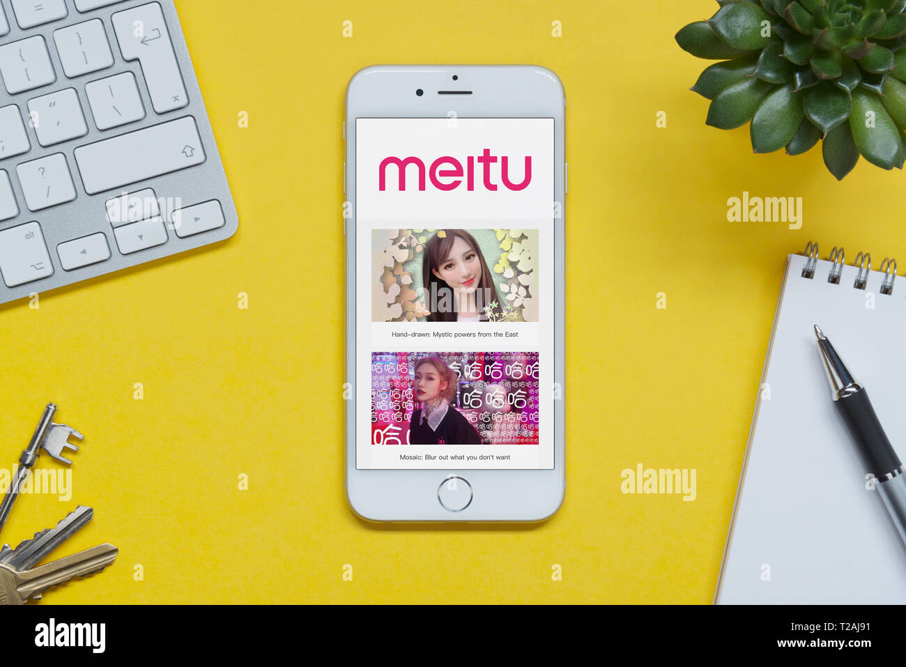 An iPhone showing the Meitu website rests on a yellow background table with a keyboard, keys, notepad and plant (Editorial use only). Stock Photo