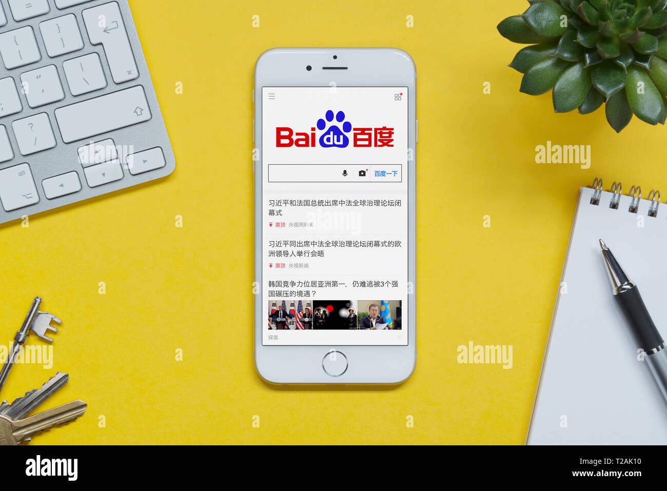 An iPhone showing the Baidu website rests on a yellow background table with a keyboard, keys, notepad and plant (Editorial use only). Stock Photo