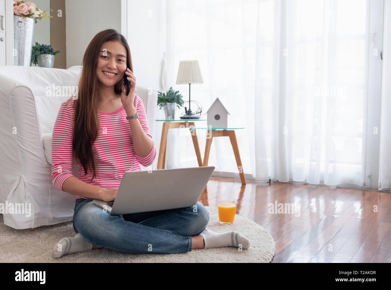 Happy asian woman talk on mobile and  typing laptop computer at home.female freelancer sitting on carpet on floor work at home in living room. Stock Photo