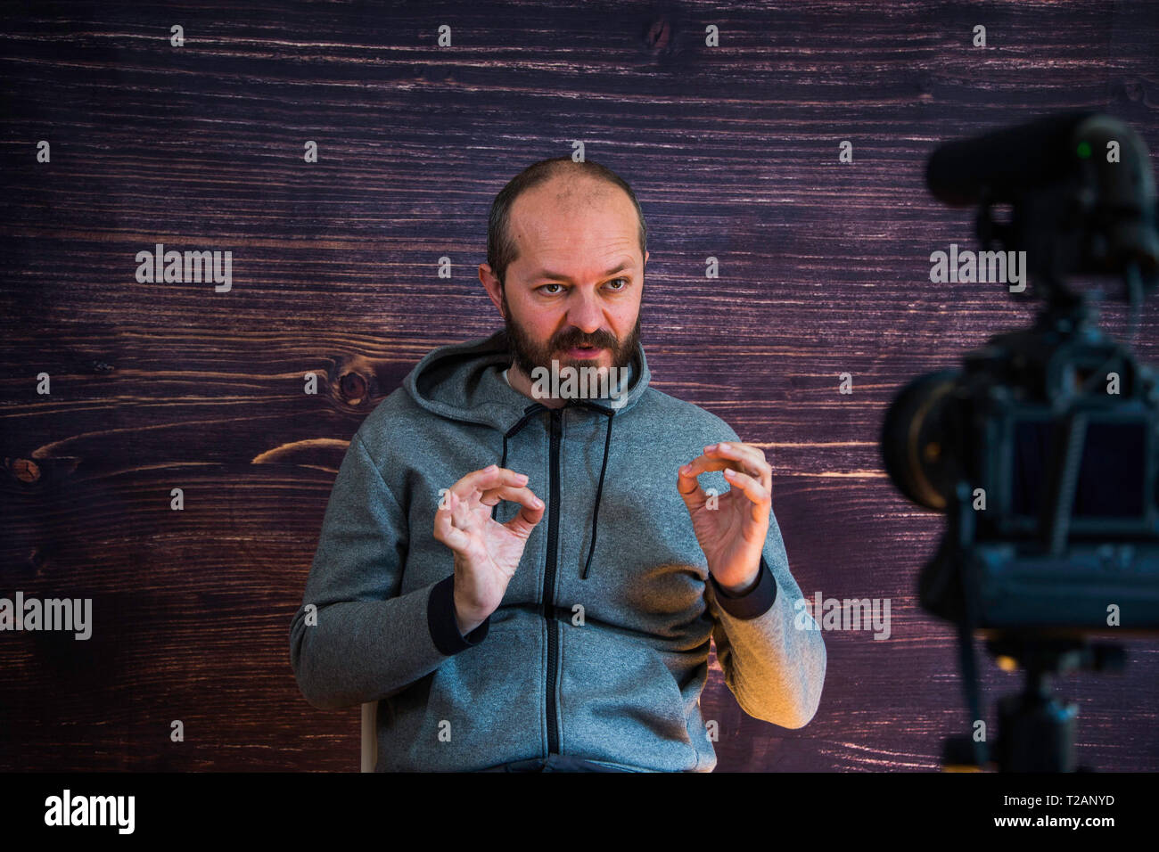 Bearded middle age man in hoody sitting and talking in front of DSLR camera, video blogging concept Stock Photo