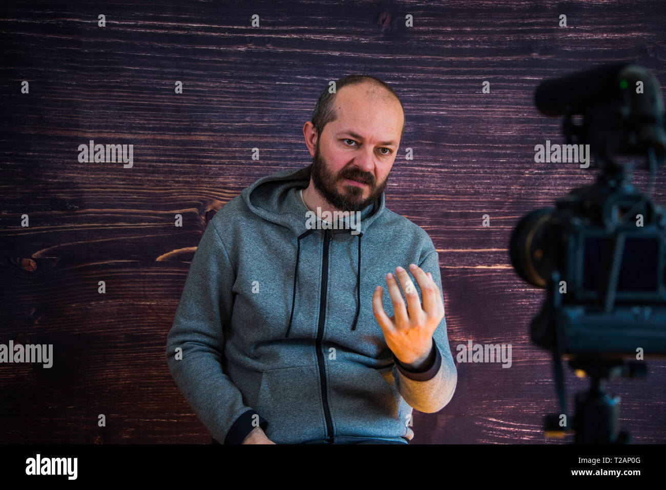 Bearded middle age man in hoody sitting and talking in front of DSLR camera, video blogging concept Stock Photo