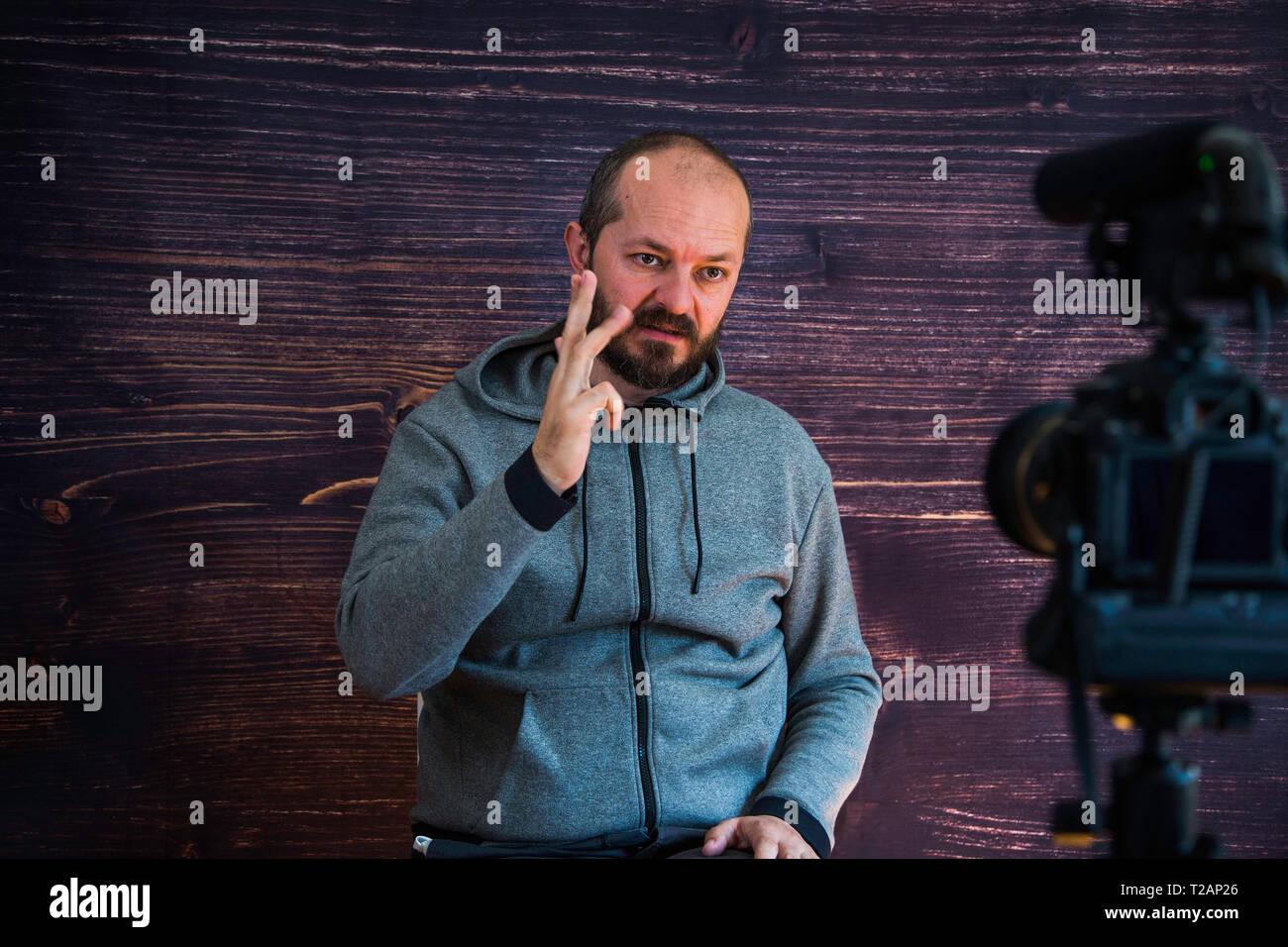 Bearded middle age man in hoody sitting and talking in front of DSLR camera, video blogging concept Stock Photo