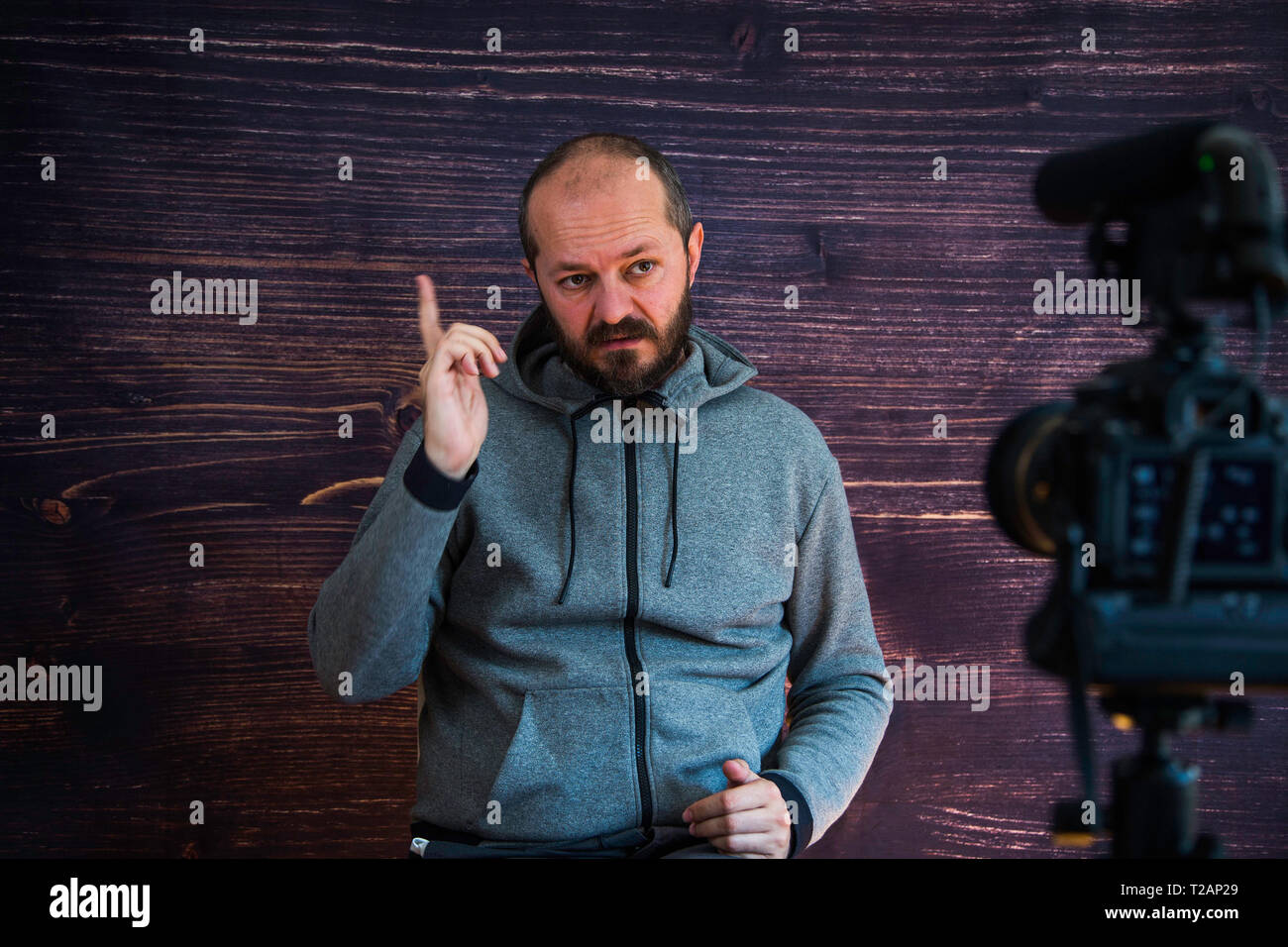 Bearded middle age man in hoody sitting and talking in front of DSLR camera, video blogging concept Stock Photo