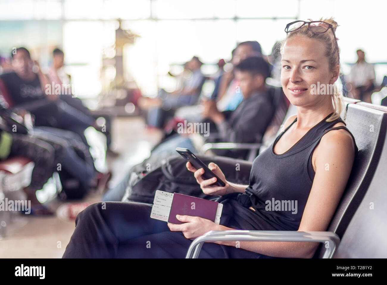 Casual tanned blond female traveler holding cell phone, passport and boarding pass while waiting to board a plane at the departure gates at the asian Stock Photo