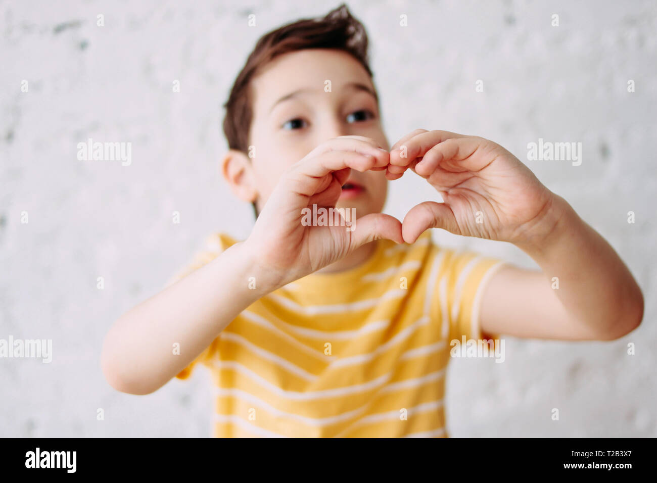 Happy tweens boy in yellow t-shirt making heart by hands on the white wall background, selective focus Stock Photo