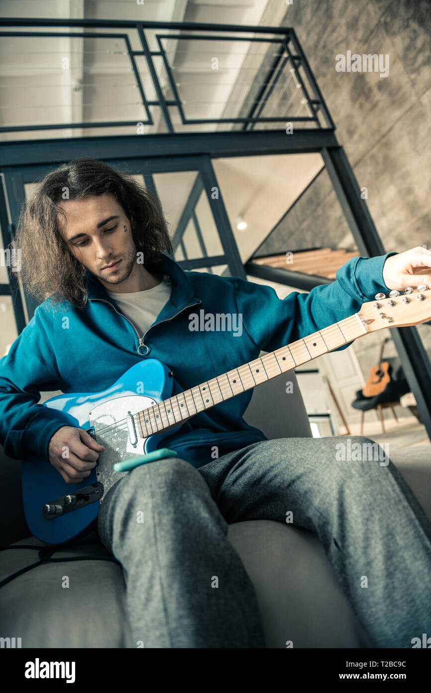 Dark-haired attentive man wearing blue sweatshirt and preparing guitar Stock Photo