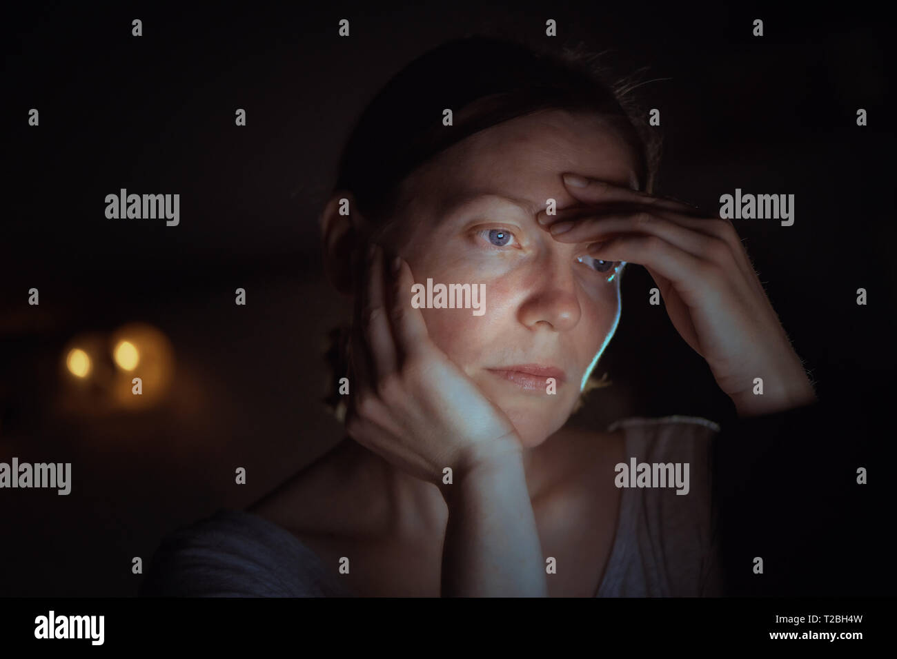 Tired woman working overtime on laptop computer at night in her home office, face is illuminated by the blue light of the screen Stock Photo