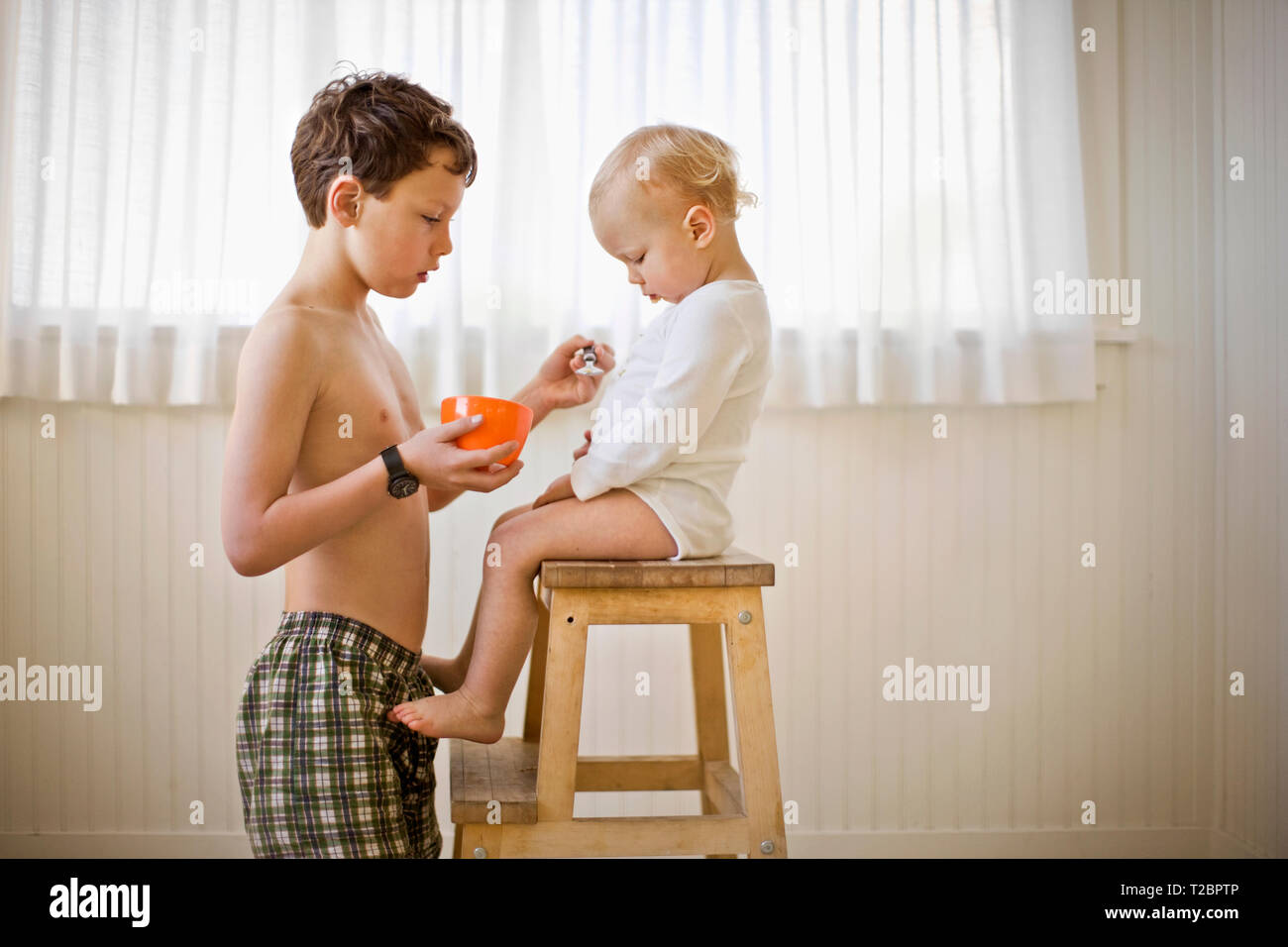 Shirtless Boy Feeding His Little Brother With A Spoon As He Sits On A