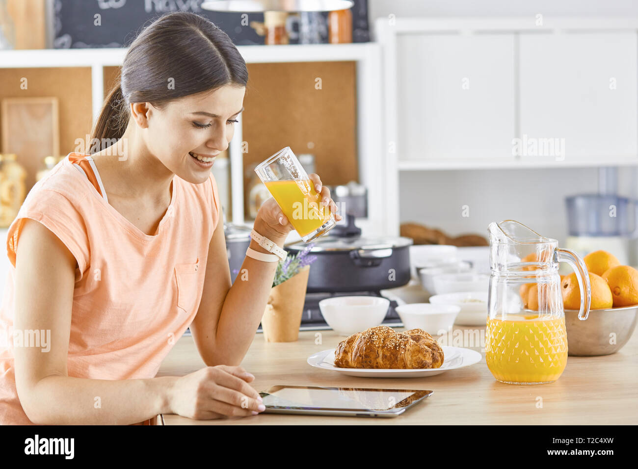 Smiling pretty woman looking at mobile phone and holding glass of orange juice while having breakfast in a kitchen. Stock Photo
