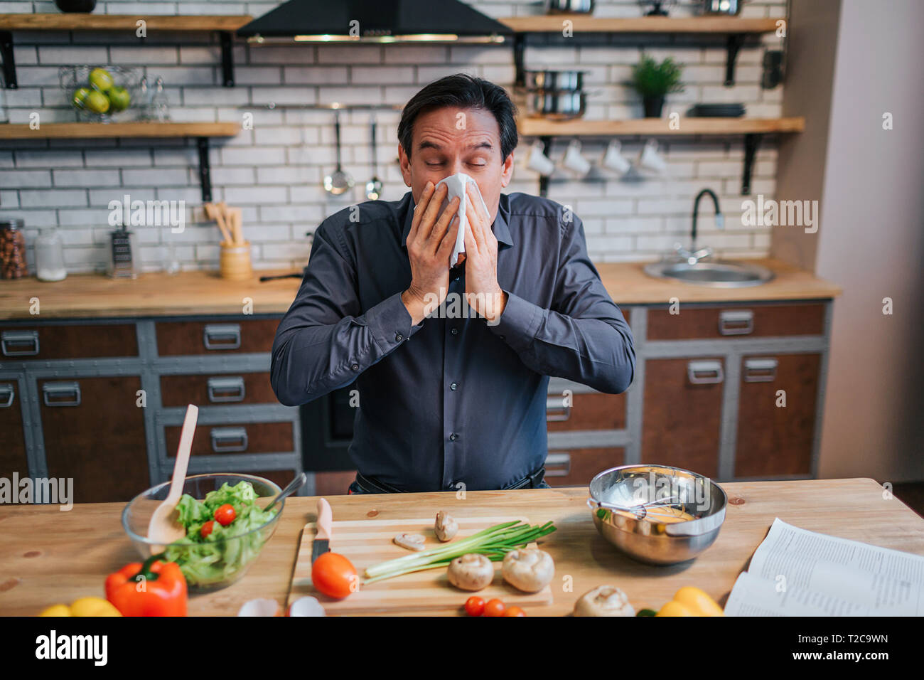 Sick adult man sneezing to white napkin. He stand at table in kitchen. Desk full of colorful healthy vegetables and spices Stock Photo