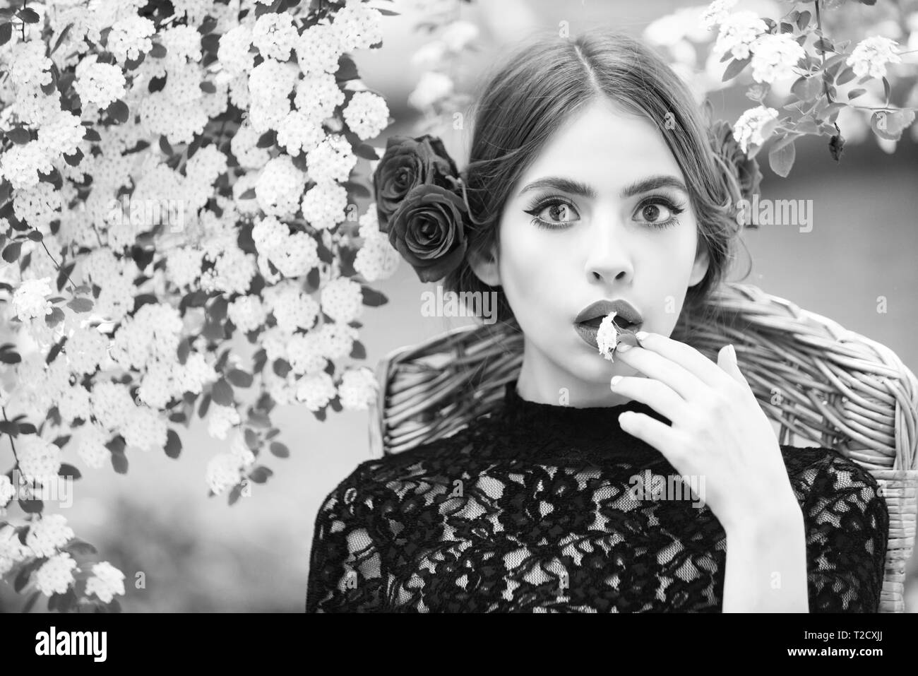 girl or cute woman with white flower in hand and stylish makeup on adorable face, with red roses in brunette hair, hairstyle, sitting on floral enviro Stock Photo