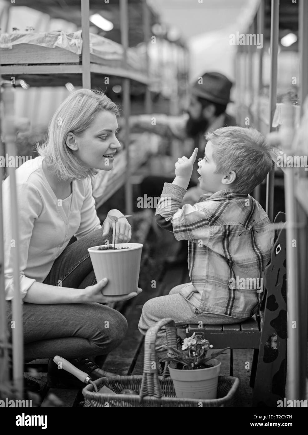 Blond kid talking to his mum while pointing up. Mother and son sitting between the rows in green house with beautiful wicker basket with gardening too Stock Photo