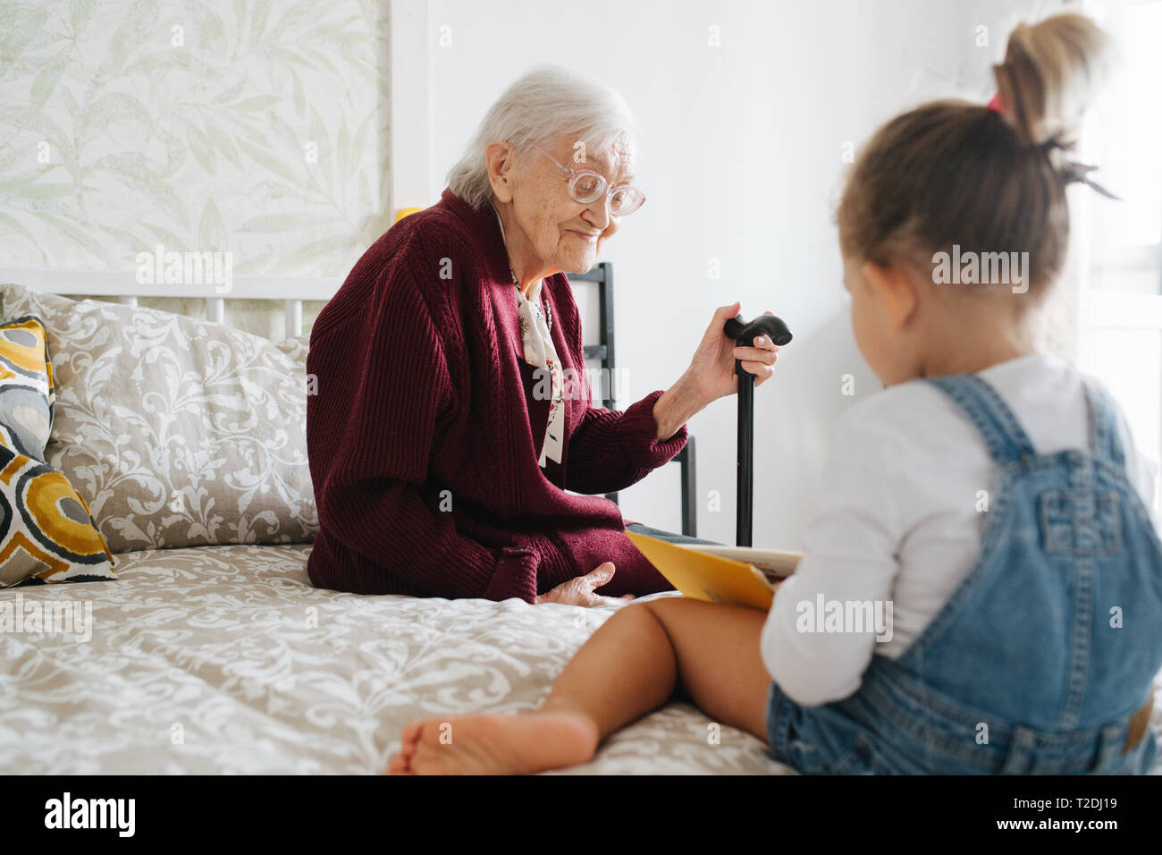 Happy moments with great grandma, senior lady spending quality time with her great granddaughter. Both sitting on a bed, girl is reading book and gran Stock Photo