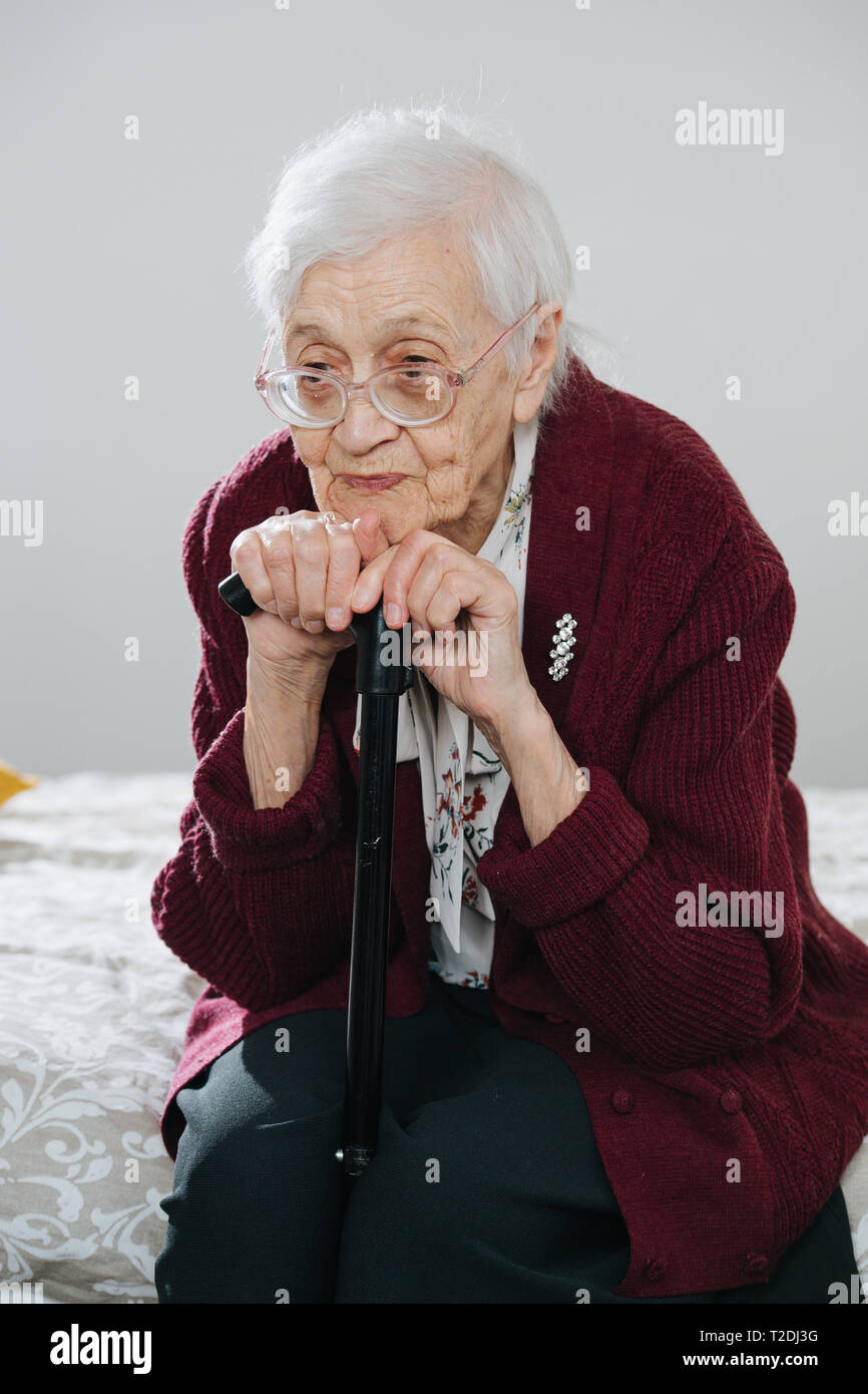 Portrait of a tired senior gray haired woman sitting on bed, leaning chin on a walking stick at home. Resting her head on a cane. Stock Photo