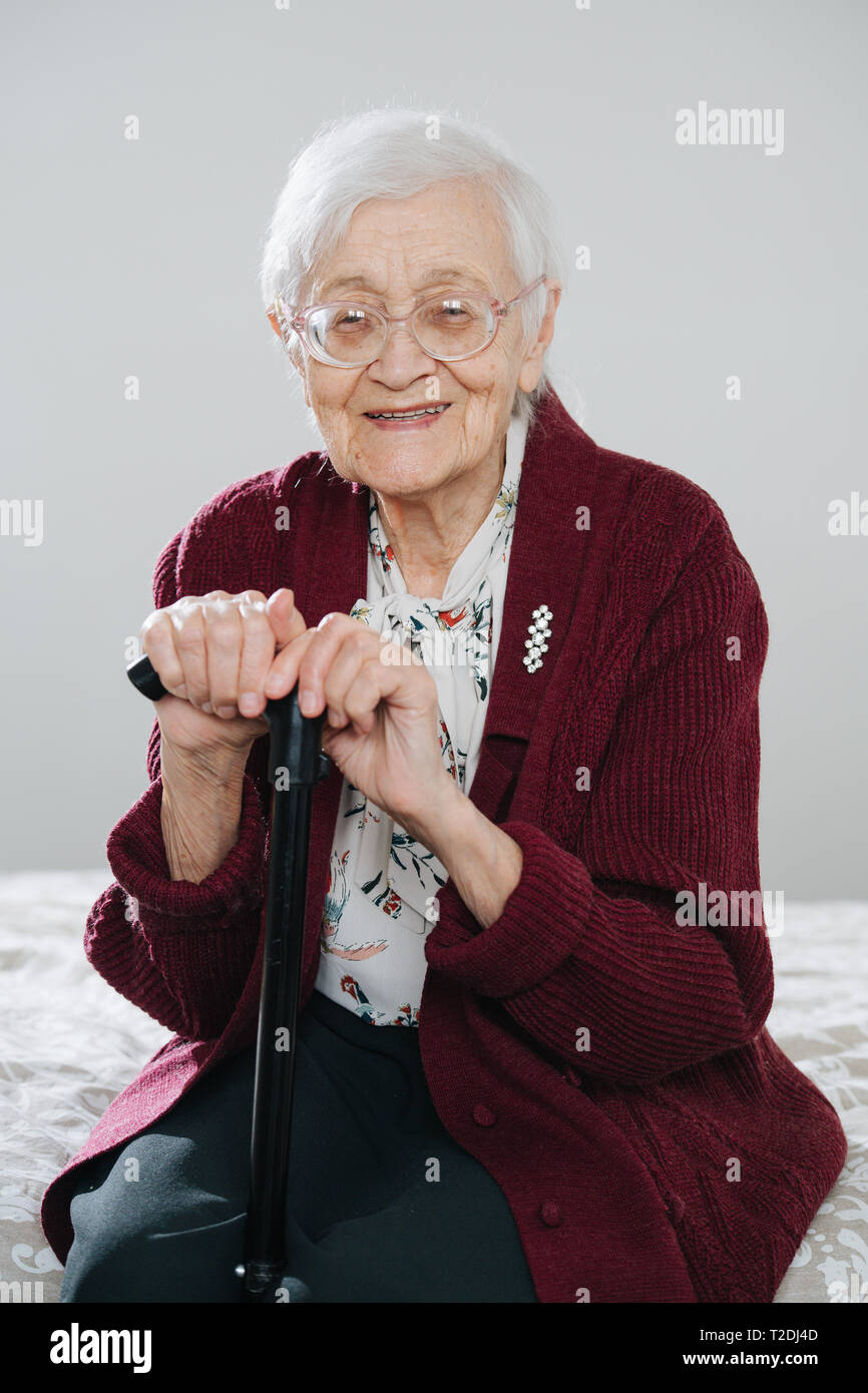 Especially cheerful senior gray haired woman sitting on bed, holding onto walking stick at home. Stock Photo