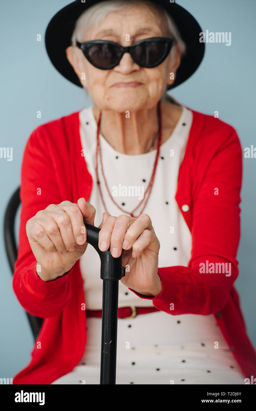 Respectable well dressed in black mini hat and sunglasses gray haired fancy senior woman with cane is resting on a chair at home on a sunny spring day Stock Photo