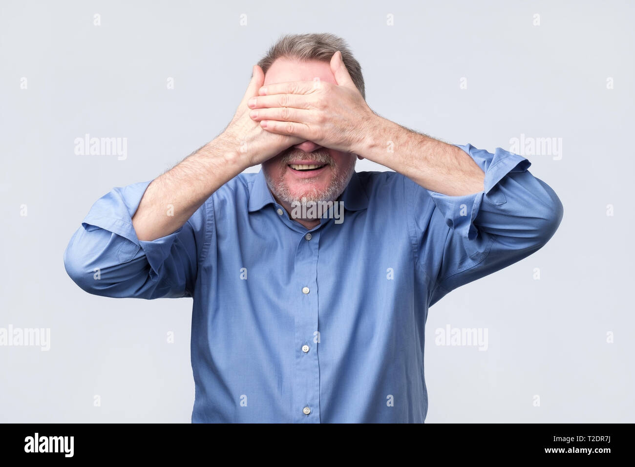 Senior happy man in blue shirt covering eyes with hands waiting for birthday surprise. Positive facial emotion. Stock Photo