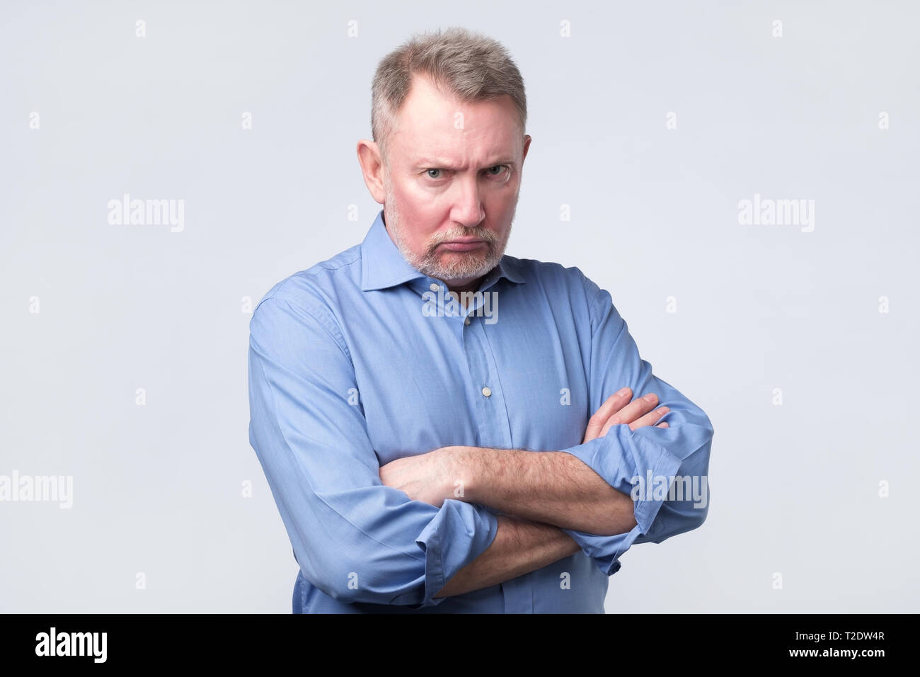 Offended senior man in blue shirt holding hands crossed on chest, looking from under forehead standing angry. Negative facial emotion Stock Photo