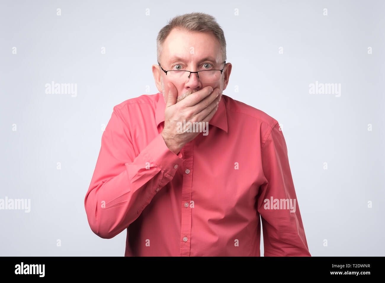 Senior man in red shirt with hand over his mouth, stunned and speechless. studio shoot. Stock Photo