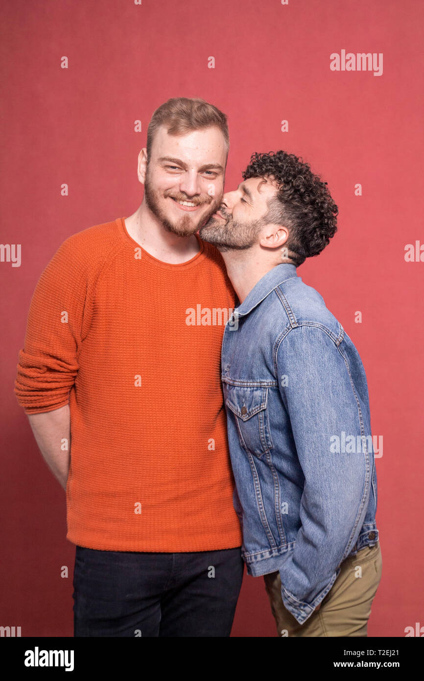 two men, gay couple posing together, smiling and happy. one is looking directly to camera. other is kissing him in the cheek. Stock Photo