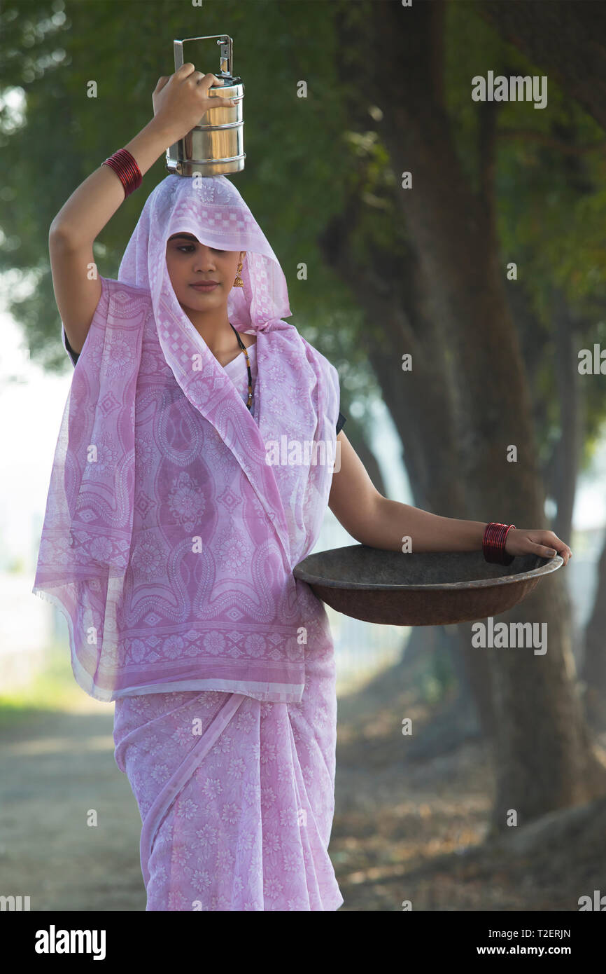 Rural woman or daily wage labourer in saree carrying an iron gold pan in hand and a tiffin box on her head to work. Stock Photo