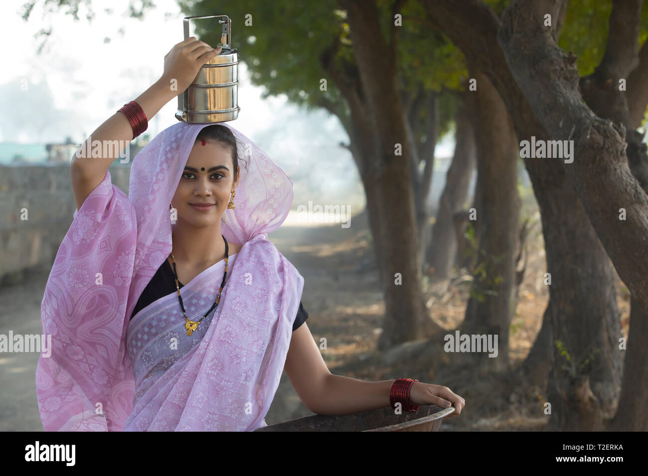 Smiling rural woman or daily wage labourer in saree carrying an iron gold pan in hand and a tiffin box on her head to work. Stock Photo