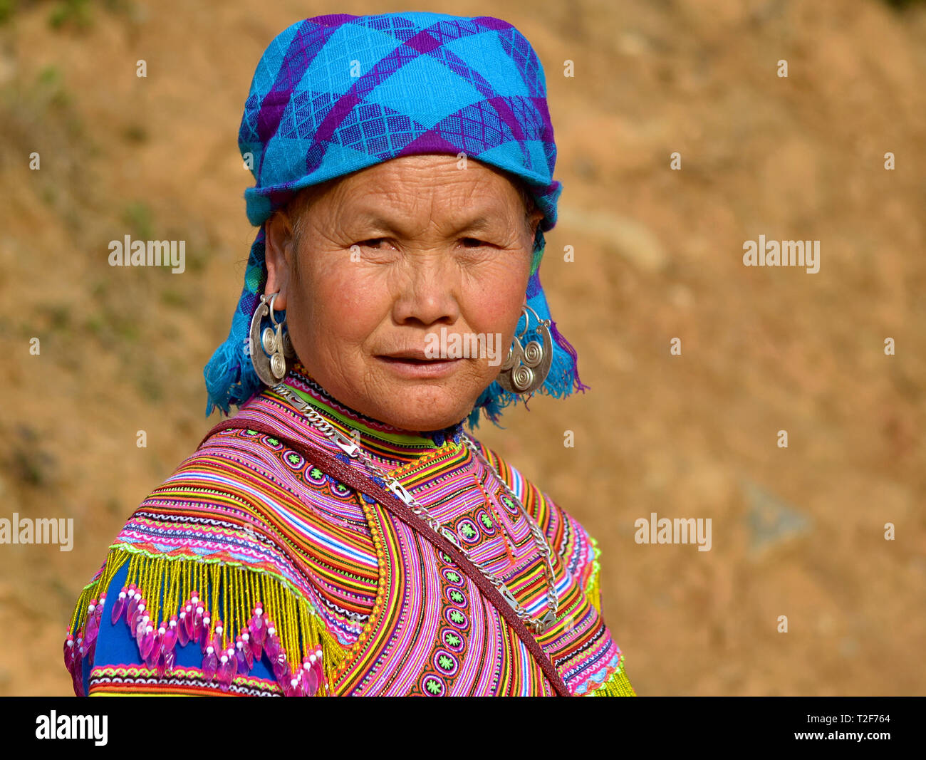 Elderly Vietnamese Flower H’mong hill-tribe market woman wears embroidered, colourful Flower H’mong traditional attire with large silver earrings. Stock Photo