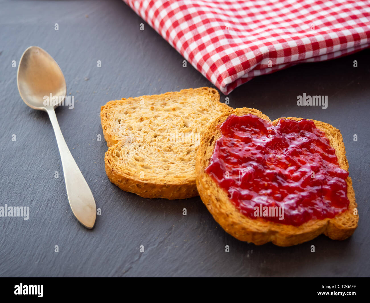Two slices of toast with raspberry jam on a slate plate, a spoon and a tablecloth old style Stock Photo